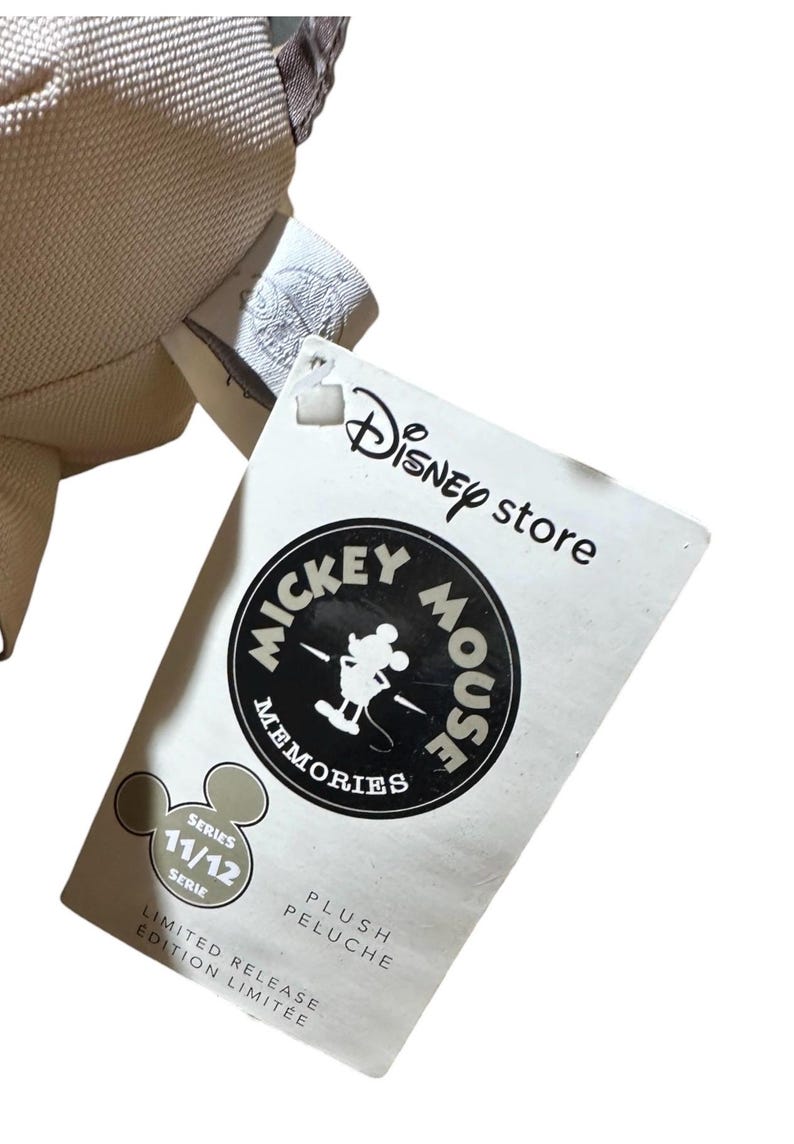 Puede incluir: Una etiqueta de Disney Store con el logotipo de Mickey Mouse Memories. La etiqueta es blanca con texto negro y un logotipo circular con Mickey Mouse. La etiqueta tambi&eacute;n incluye el texto "Serie 11/12" y "Edici&oacute;n Limitada".