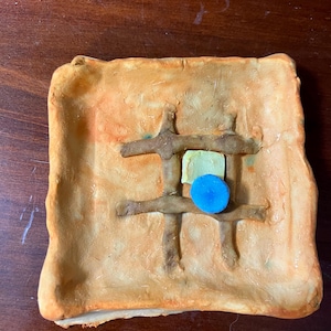 May include: A square, terracotta-colored ceramic dish with a raised, cross-shaped design. The dish has a light blue and green circular object in the center.
