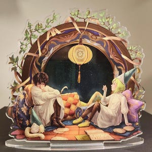 Puede incluir: Soporte decorativo de acrílico que presenta dos figuras con túnicas blancas, sentadas dentro de un arco estilizado. La escena incluye una linterna colgante, almohadas y un fondo estrellado. El soporte tiene una base transparente.