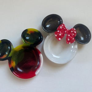 以下が含まれることがあります： ミッキーマウスとミニーマウスの形をした2つの装飾的な樹脂皿。1つは白い皿で、黒い耳と白い水玉模様の赤いリボンが付いています。もう1つは赤、黄、緑、黒が混ざり合ったもので、お揃いの耳が付いています。