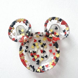 Puede incluir: Cenicero de resina transparente con forma de la cabeza de Mickey Mouse. El cenicero está lleno de confeti colorido con forma de Mickey Mouse en rojo, negro, amarillo y blanco. Las orejas también contienen confeti.