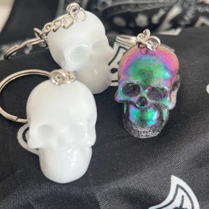 Puede incluir: Tres llaveros con forma de calavera. Dos son blancos y uno tiene un acabado brillante e iridiscente con tonos rosas, verdes y azules. Cada llavero tiene un anillo y una cadena de color plateado. Los llaveros están sobre una superficie negra estampada.