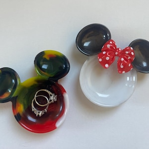 Könnte beinhalten: Zwei Mickey Mouse-förmige Ringhalter. Einer ist ein farbenfroher Wirbel aus Rot, Gelb und Grün mit zwei Ringen. Der andere ist weiß mit schwarzen Ohren und einer roten Schleife mit weißen Punkten. Eine unterhaltsame und dekorative Möglichkeit, Schmuck aufzubewahren.