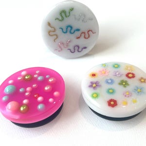 May include: Three decorative magnets with colorful designs. One white magnet features glittery snakes in various colors. A pink magnet has assorted pastel-colored beads. A white magnet displays colorful flowers.