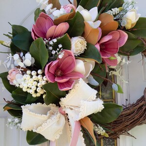 May include: A decorative wreath featuring a natural grapevine base adorned with faux magnolia blooms in shades of pink, white, and orange. The wreath is accented with green leaves, white berries, and a cream-colored bow with lace trim.