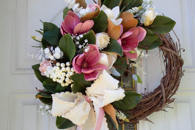 May include: A decorative wreath featuring a natural grapevine base adorned with faux magnolia blooms in shades of pink, white, and orange. The wreath is accented with green leaves, white berries, and a cream-colored bow with lace trim.