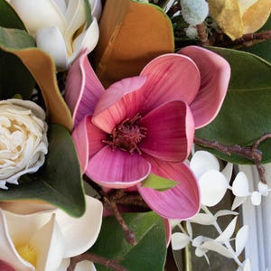 May include: Close-up of a floral arrangement featuring large, pink magnolia blossoms with dark centers. The arrangement includes white and cream-colored flowers, green leaves, and brown stems, creating a natural, textured aesthetic.