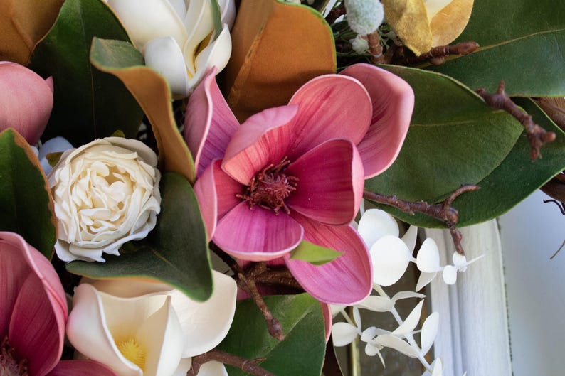 May include: Close-up of a floral arrangement featuring large, pink magnolia blossoms with dark centers. The arrangement includes white and cream-colored flowers, green leaves, and brown stems, creating a natural, textured aesthetic.