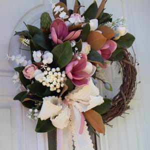 May include: A magnolia wreath with a grapevine base, featuring large pink and white magnolia blooms, green leaves, and white berries. A cream-colored ribbon with lace trim and a pink ribbon accent the wreath, suitable for a front door.