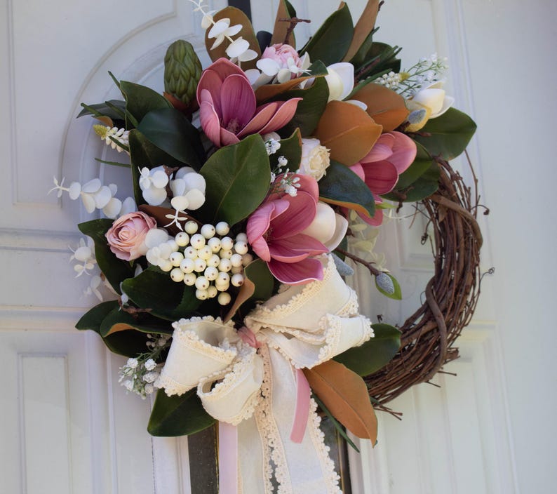 May include: A magnolia wreath with a grapevine base, featuring large pink and white magnolia blooms, green leaves, and white berries. A cream-colored ribbon with lace trim and a pink ribbon accent the wreath, suitable for a front door.