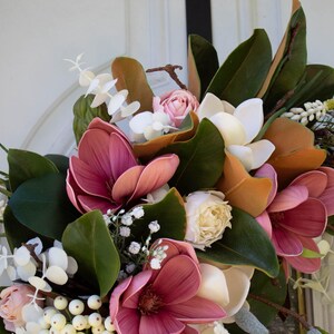 May include: A close-up of a floral wreath featuring large, deep pink magnolia blooms, cream-colored roses, and white accents. The wreath includes green leaves and brown branches, creating a natural, elegant design.