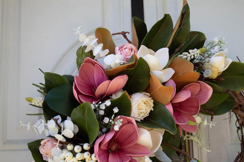 May include: A close-up of a floral wreath featuring large, deep pink magnolia blooms, cream-colored roses, and white accents. The wreath includes green leaves and brown branches, creating a natural, elegant design.