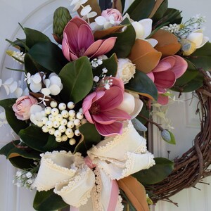May include: A decorative wreath featuring a variety of faux flowers and greenery. The wreath includes large, deep pink magnolia blooms, white berries, and a cream-colored ribbon bow. The base is made of woven brown twigs.