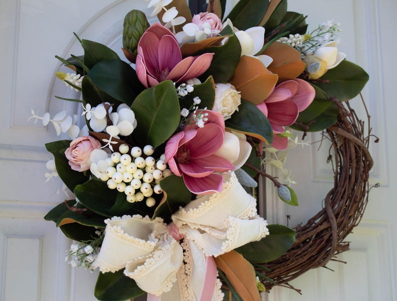 May include: A decorative wreath featuring a variety of faux flowers and greenery. The wreath includes large, deep pink magnolia blooms, white berries, and a cream-colored ribbon bow. The base is made of woven brown twigs.