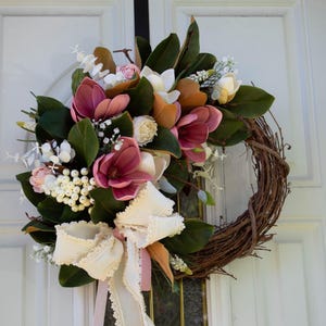 May include: A handcrafted wreath featuring a grapevine base adorned with faux magnolia blooms in shades of pink, white, and orange. The wreath is accented with green leaves, white berries, and a cream-colored bow with pink ribbon, perfect for seasonal home decor.
