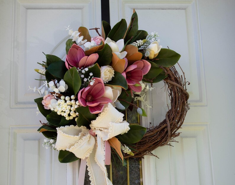 May include: A handcrafted wreath featuring a grapevine base adorned with faux magnolia blooms in shades of pink, white, and orange. The wreath is accented with green leaves, white berries, and a cream-colored bow with pink ribbon, perfect for seasonal home decor.