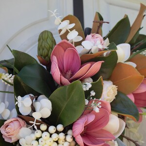 May include: A floral wreath featuring pink magnolia blooms, green leaves, and white berries. The wreath includes white and yellow accent flowers and brown leaves. The wreath is set against a white background.