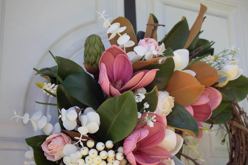 May include: A floral wreath featuring pink magnolia blooms, green leaves, and white berries. The wreath includes white and yellow accent flowers and brown leaves. The wreath is set against a white background.