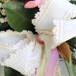 May include: Close-up of a floral wreath with a large white bow. The bow is made of white fabric with a delicate lace trim. The wreath also features green leaves, white berries, and pink and white flowers.