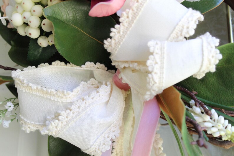 May include: Close-up of a floral wreath with a large white bow. The bow is made of white fabric with a delicate lace trim. The wreath also features green leaves, white berries, and pink and white flowers.