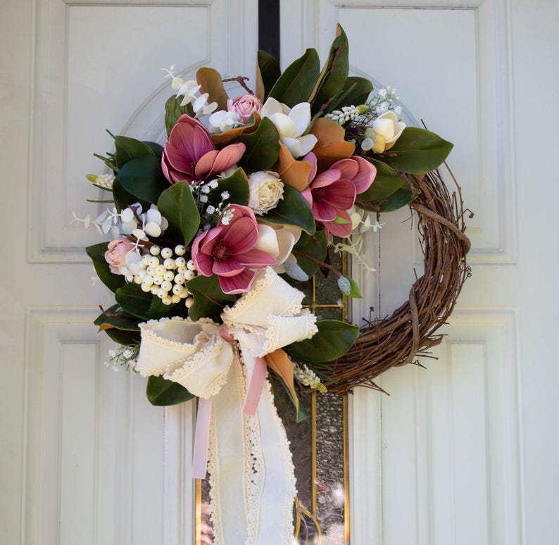 May include: A handcrafted wreath featuring a grapevine base adorned with faux magnolia blooms in shades of pink, white, and orange. The wreath is accented with green leaves, white berries, and a cream-colored bow with lace trim. A decorative piece for the front door.