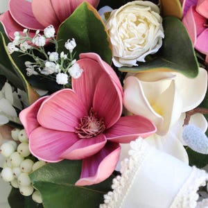 May include: A close-up of a floral wreath featuring large pink magnolia blooms, white roses, and small white berries. The wreath includes green leaves and a white ribbon with lace trim, creating a natural and elegant design.