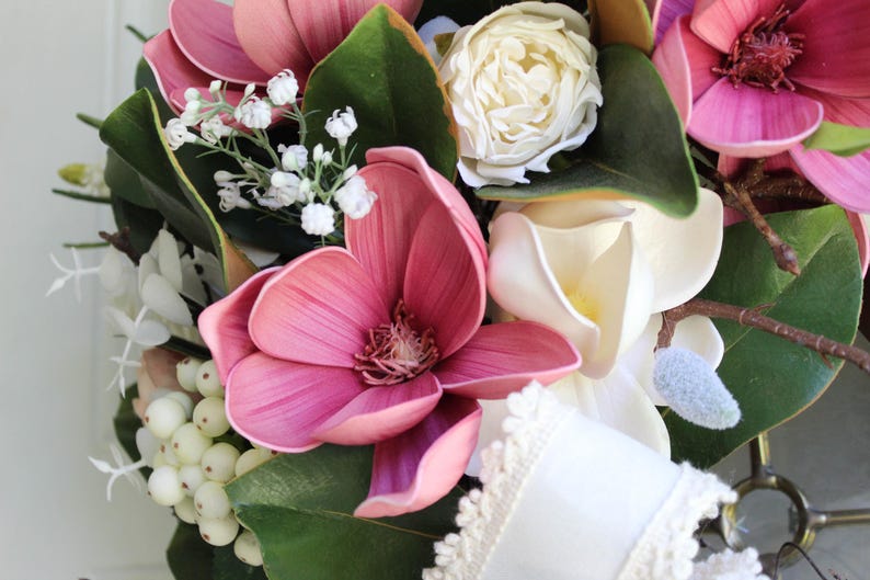 May include: A close-up of a floral wreath featuring large pink magnolia blooms, white roses, and small white berries. The wreath includes green leaves and a white ribbon with lace trim, creating a natural and elegant design.