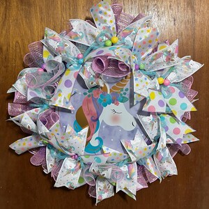 May include: A whimsical unicorn wreath featuring a central image of a unicorn with a floral crown. The wreath is adorned with white ribbons patterned with unicorns, stars, and polka dots, and accented with pink and purple mesh.