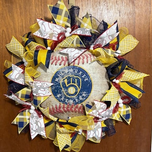 May include: A yellow, blue, and red Milwaukee Brewers baseball wreath with a baseball-themed background and white baseballs. The wreath is decorated with yellow, blue, and red ribbons and white beads.