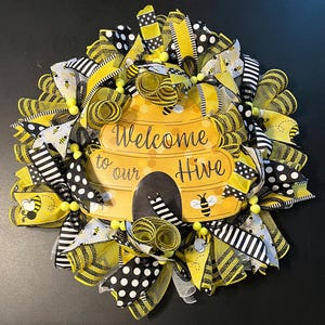 May include: A decorative wreath featuring a yellow and black beehive design with the text "Welcome to our Hive." The wreath is adorned with ribbons in yellow, black, and white patterns, including stripes and polka dots, and small bee accents.