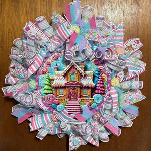 May include: A decorative wreath featuring a central circular sign depicting a gingerbread house with candy accents. The wreath is constructed with layers of mesh ribbon in pink, blue, and white, accented with candy-themed patterns and glitter.