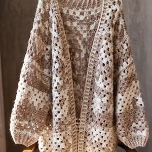 May include: A handmade, open-front crochet cardigan in a neutral color palette of cream and brown. The oversized sweater features a textured, open stitch design and long sleeves. The cardigan is displayed on a wooden chair.