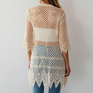 May include: A cream-colored, open-weave crochet cardigan with three-quarter sleeves. The cardigan has a scalloped hem and a loose, flowing design. The back of the cardigan is visible, showing a white top and blue jeans underneath. The crochet pattern is intricate and detailed.