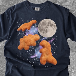 May include: Navy blue t-shirt featuring a whimsical design of dinosaur-shaped chicken nuggets floating in a starry galaxy with a full moon. The nuggets are golden brown, and the moon is detailed. A fun, playful graphic tee.