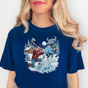 Puede incluir: Camiseta azul marino con un estampado gráfico. El gráfico representa a un leñador cortando hielo con un hacha, un toro azul y una cabaña en un bosque nevado. El leñador lleva una camisa a cuadros roja y negra.