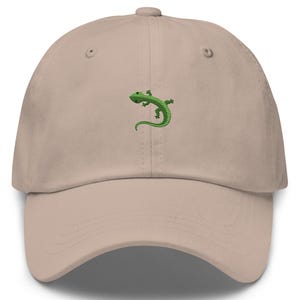 Può includere: Cappellino da baseball beige con un'emblema di lucertola verde ricamata. Il cappellino ha una visiera curva e un classico design a sei pannelli. Presenta un bottone sulla corona e un cinturino regolabile sul retro.