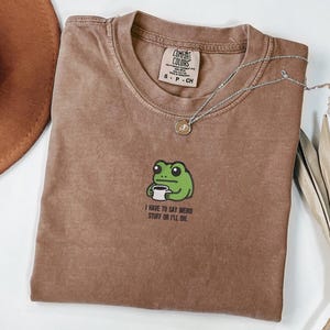 Può includere: T-shirt marrone con girocollo e grafica di una rana verde che tiene una tazza di caffè. Il testo sotto la rana recita "I HAVE TO SAY WEIRD STUFF OR I'LL DIE." Sono visibili anche una collana d'argento e un cappello marrone.