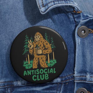 Puede incluir: Un botón redondo negro con un dibujo de Bigfoot sosteniendo una bebida y haciendo el signo de la paz. La imagen incluye árboles verdes y el texto "ANTISOCIAL CLUB" en verde.