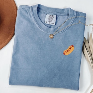 May include: A blue Comfort Colors t-shirt with a small embroidered hot dog. The shirt has a crew neck and short sleeves. A gold necklace with a pendant is draped across the shirt. A brown hat is partially visible in the upper left corner.