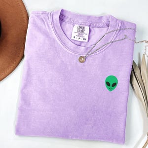 Op de afbeelding: Een lavendelkleurig Comfort Colors T-shirt met een klein groen aliengezichtje geborduurd op de voorkant. Een zilveren ketting met een hanger ligt op het shirt. Het gevouwen T-shirt wordt op een wit oppervlak gepresenteerd.
