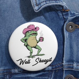 May include: A white button featuring a cartoon frog wearing a pink cowboy hat, holding a pink mug with steam rising. The frog has a grumpy expression. The text "Well Sheeeyit" is written in a cursive font below the frog.