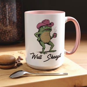 May include: White ceramic mug with a pink interior and handle. The mug features a cartoon frog wearing a cowboy hat and holding a coffee cup. The text "Well Sheeeyit" is written below the frog.