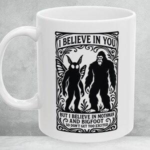 May include: White ceramic mug with a black graphic featuring Mothman and Bigfoot, with the text "I BELIEVE IN YOU BUT I BELIEVE IN MOTHMAN AND BIGFOOT SO DON'T GET TOO EXCITED". The mug has a curved handle.