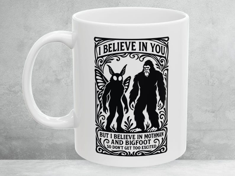 May include: White ceramic mug with a black graphic featuring Mothman and Bigfoot, with the text "I BELIEVE IN YOU BUT I BELIEVE IN MOTHMAN AND BIGFOOT SO DON'T GET TOO EXCITED". The mug has a curved handle.