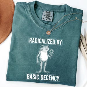 May include: A teal Comfort Colors t-shirt with the text "RADICALIZED BY BASIC DECENCY" and a graphic of a frog holding a flower. A gold necklace and a brown hat are also in the image.