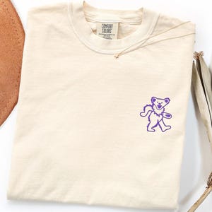 May include: A cream-coloured short-sleeve t-shirt featuring a small purple graphic of a dancing bear on the left chest. The inner neck label displays the text Comfort Colors, indicating the brand and garment details in a clear, readable font.