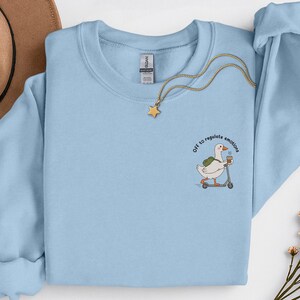 May include: Pale blue crew neck sweatshirt. The design features a goose on a scooter, carrying a coffee cup, with the text "Off to regulate emotions". A gold necklace with a star charm is draped across the sweatshirt.