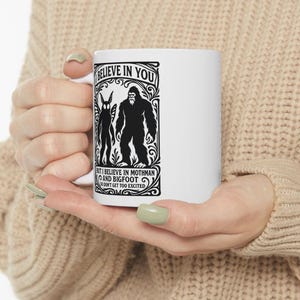 May include: White ceramic mug featuring a black graphic of Mothman and Bigfoot, with the text "I BELIEVE IN YOU" and "BUT I BELIEVE IN MOTHMAN AND BIGFOOT DON'T GET TOO EXCITED". The mug is held in a person's hands.