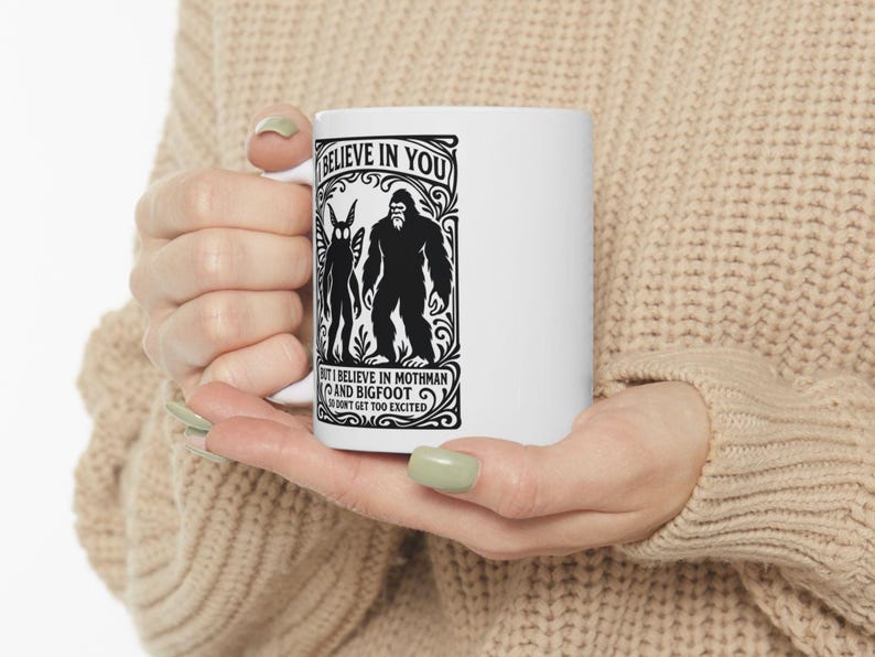 May include: White ceramic mug featuring a black graphic of Mothman and Bigfoot, with the text "I BELIEVE IN YOU" and "BUT I BELIEVE IN MOTHMAN AND BIGFOOT DON'T GET TOO EXCITED". The mug is held in a person's hands.