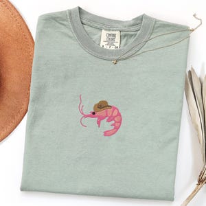 May include: A pale green t-shirt with an embroidered pink shrimp wearing a brown cowboy hat. A brown cowboy hat and a gold necklace are visible. The shirt has a Comfort Colors tag.